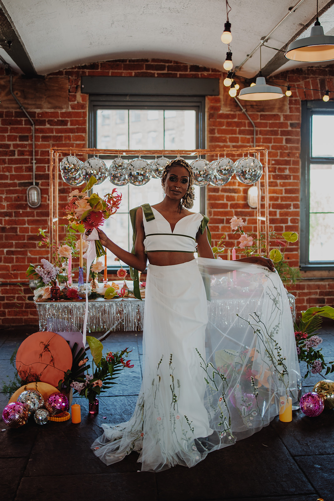 bride wearing meadow two piece wedding separates - embroidered with green floral motifs. She stands in front of alternative wedding breakfast table with disco balls.
