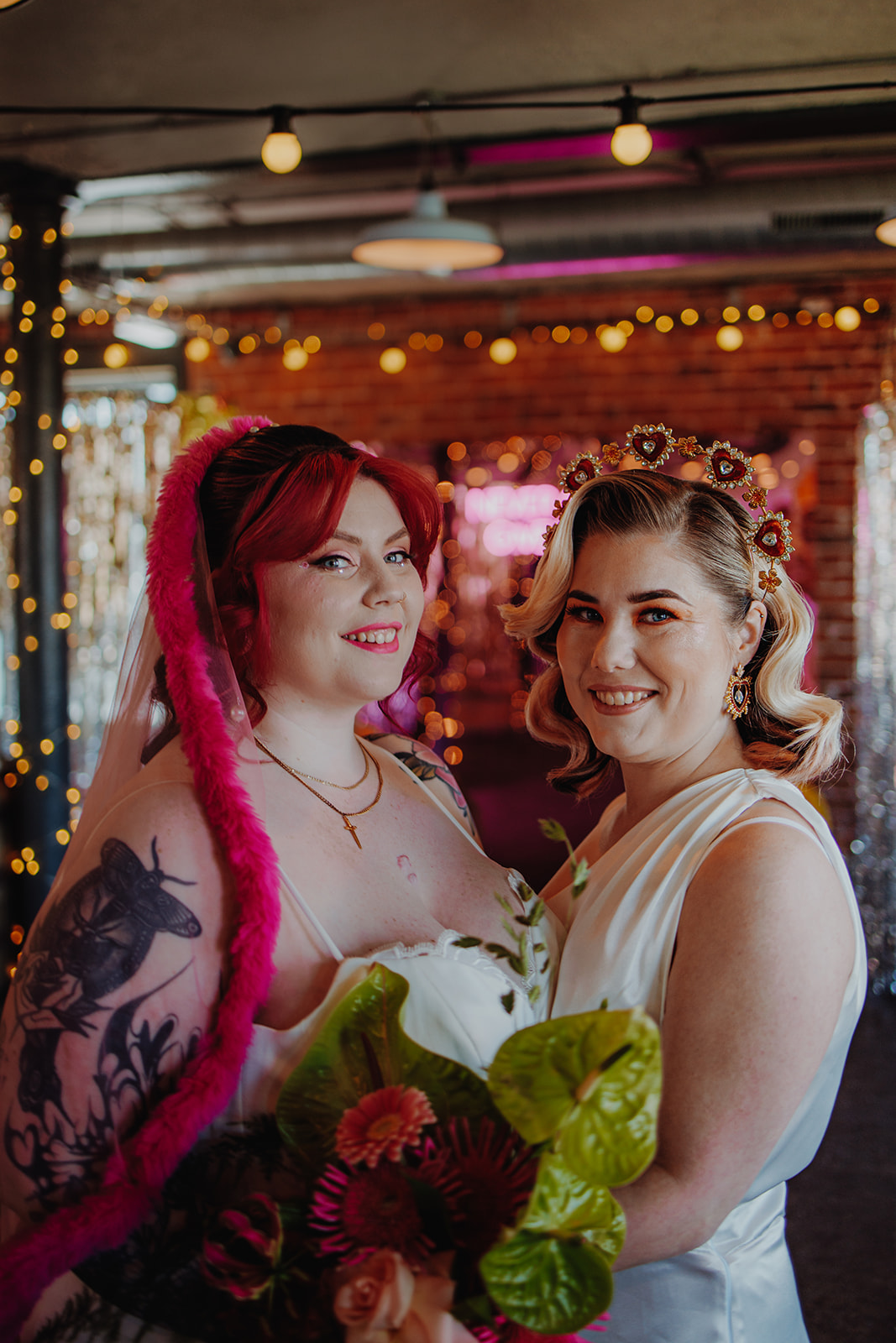 Two brides hug looking at hte camera. One wears a bright pink veil and the other a heart halo crown with hollywood waves