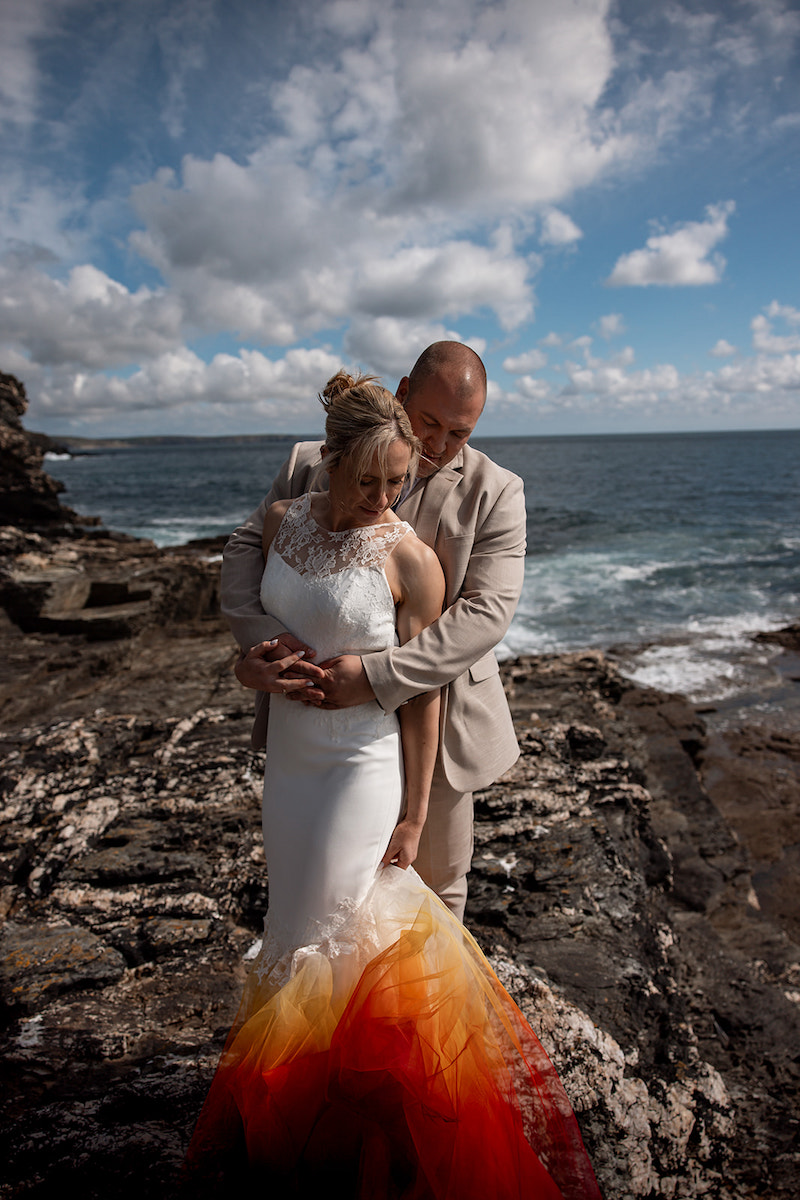 groom embracing bride wearing bright dip dye mermaid wedding dress on the Cornish coastline rocks