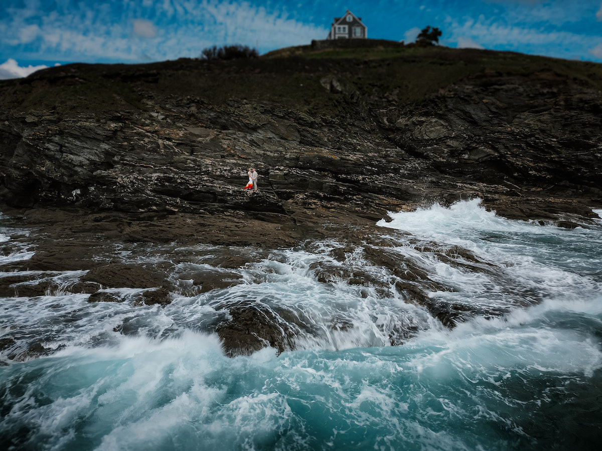 striking coastal wedding portrait where waves crash on the rocks where the couple stand. Their wedding venue is visible on the clifftop.