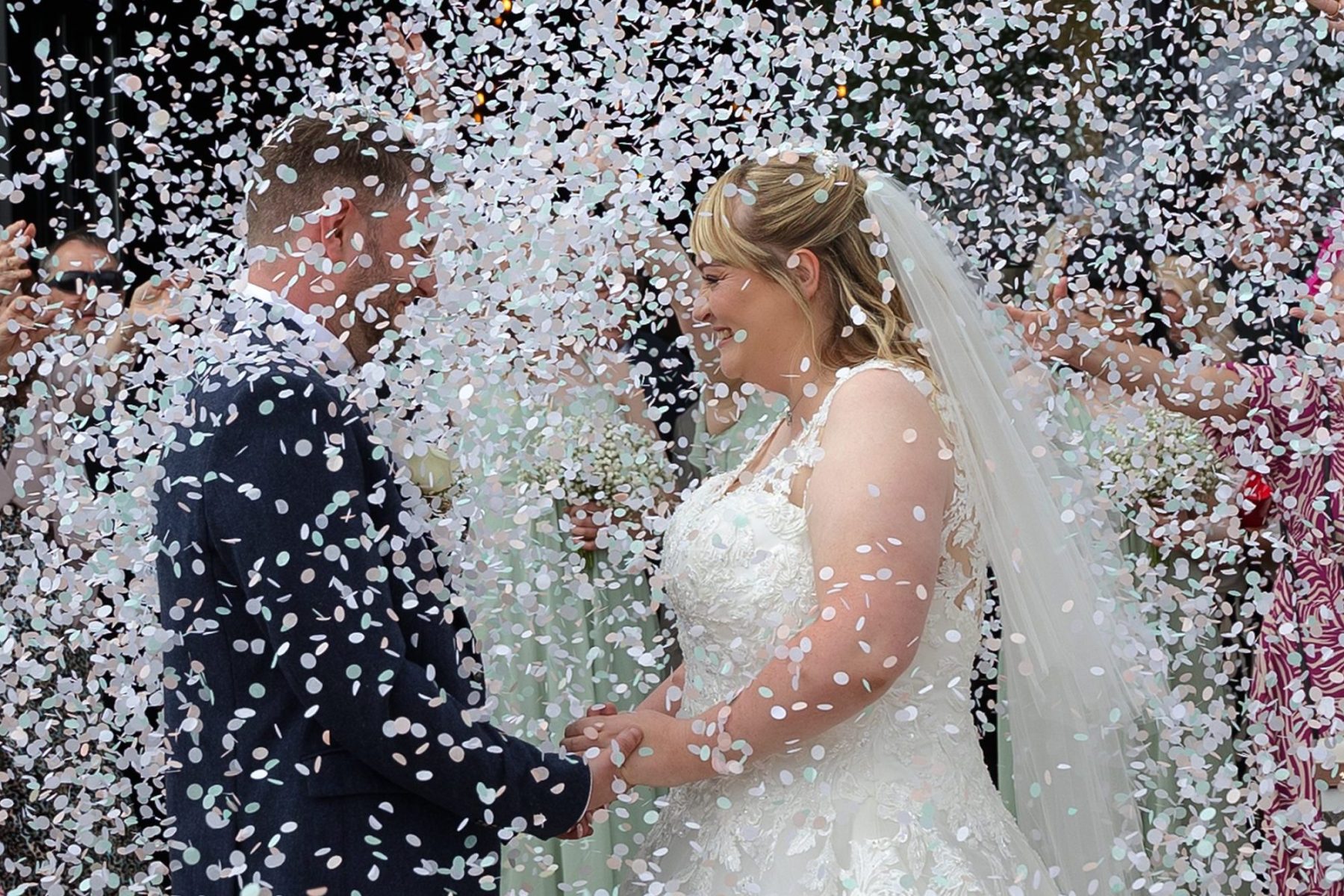 Bride and groom hold hands under a shower of white, pale pink and pale blue biodegradable wedding confetti
