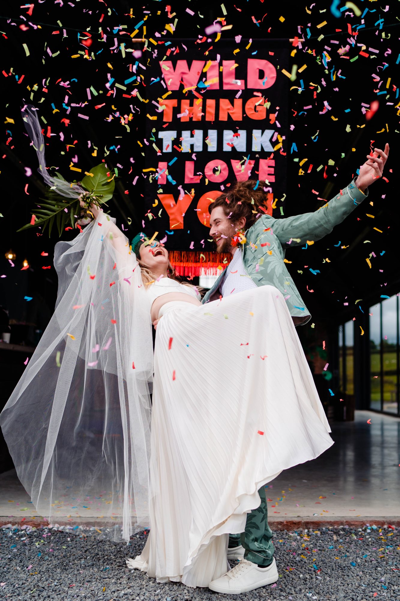 edgy newly weds pose under ticket tape style wedding confetti with 'wild thing I think I love you' banner behind