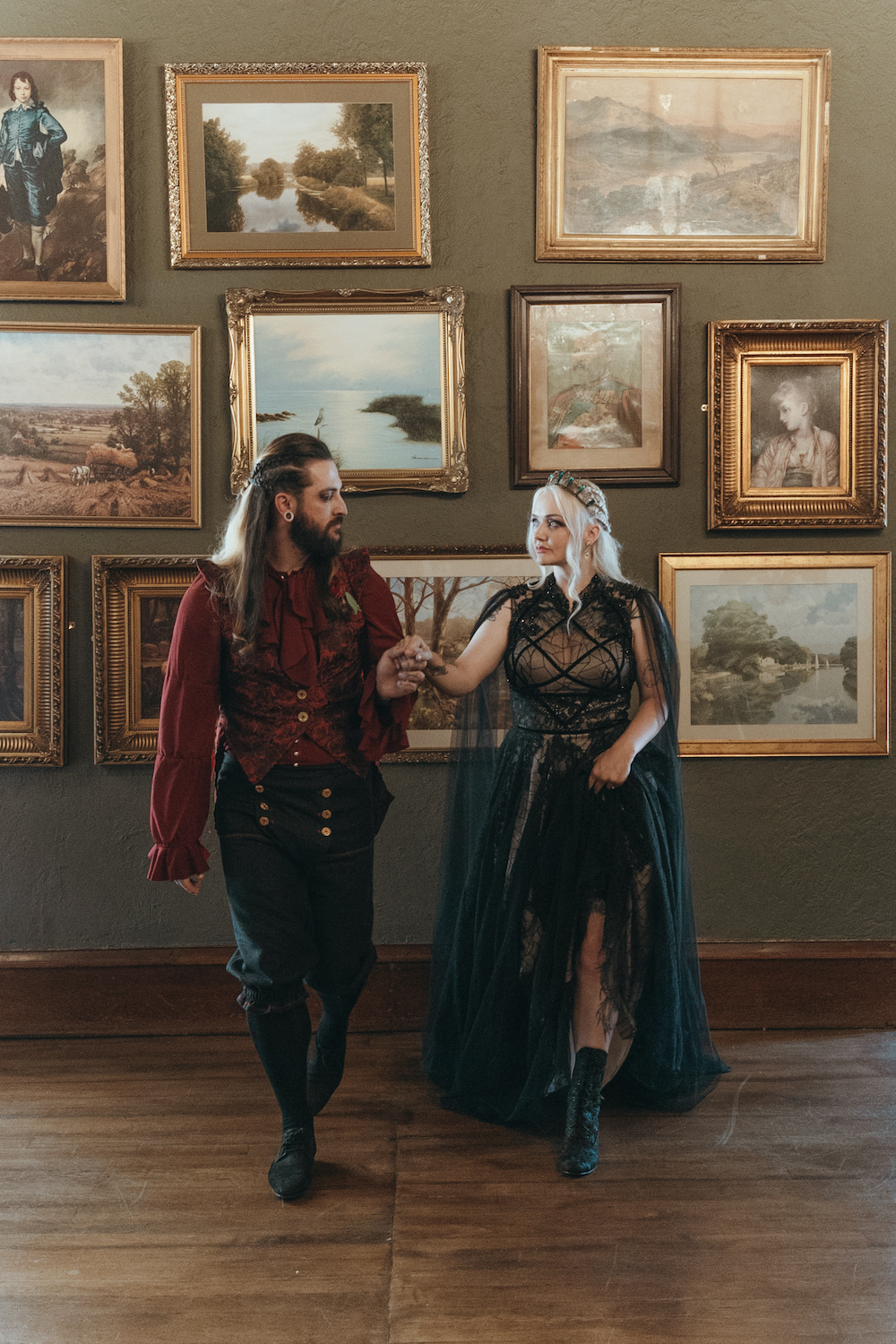 gothic fantasy bride and groom walk hand in hand through room filled with antique pictures. Bride lifts her dress to show her victorian ankle boots