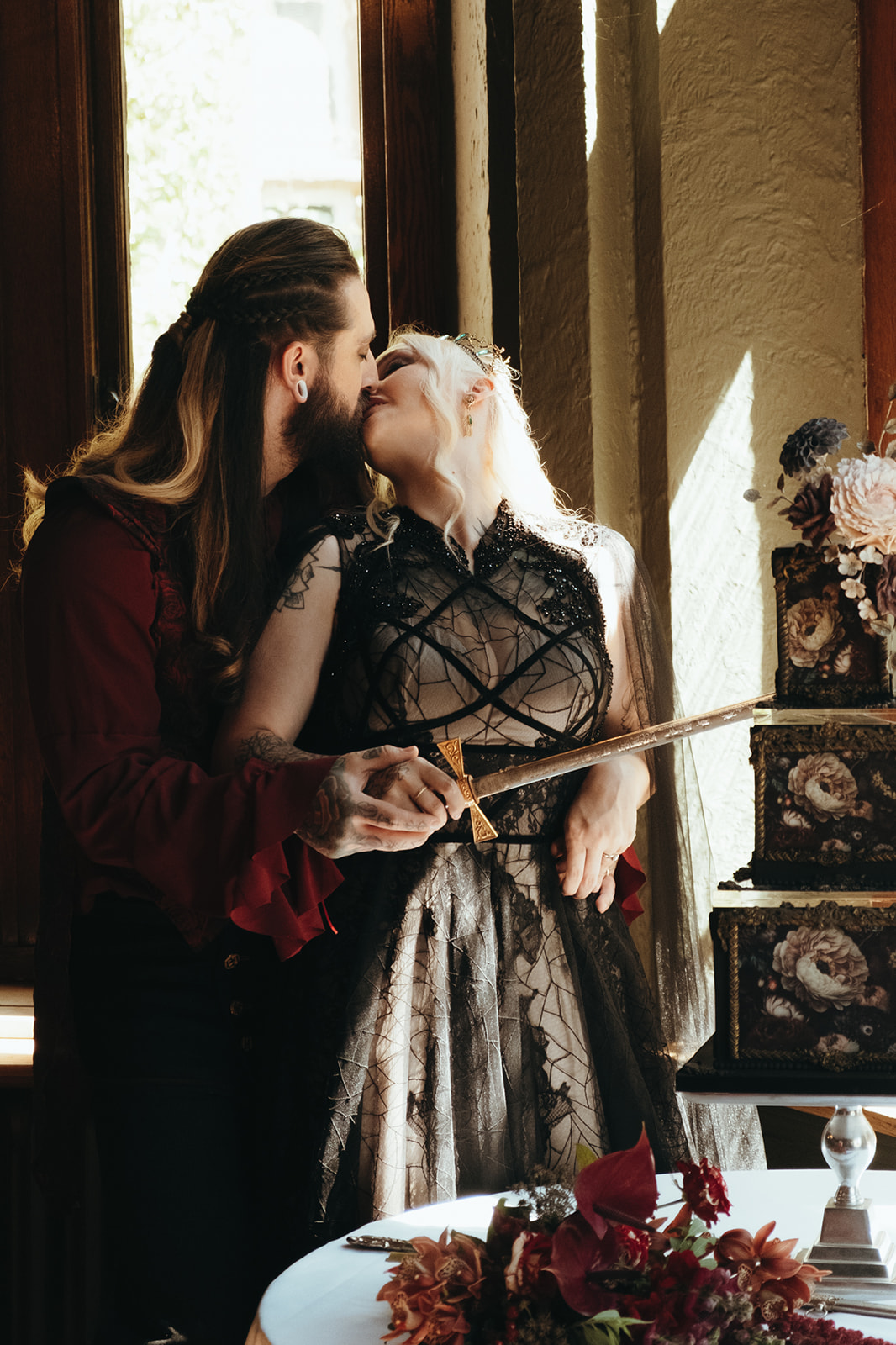 bride and groom kiss while cutting the gothic wedding cake with a sword