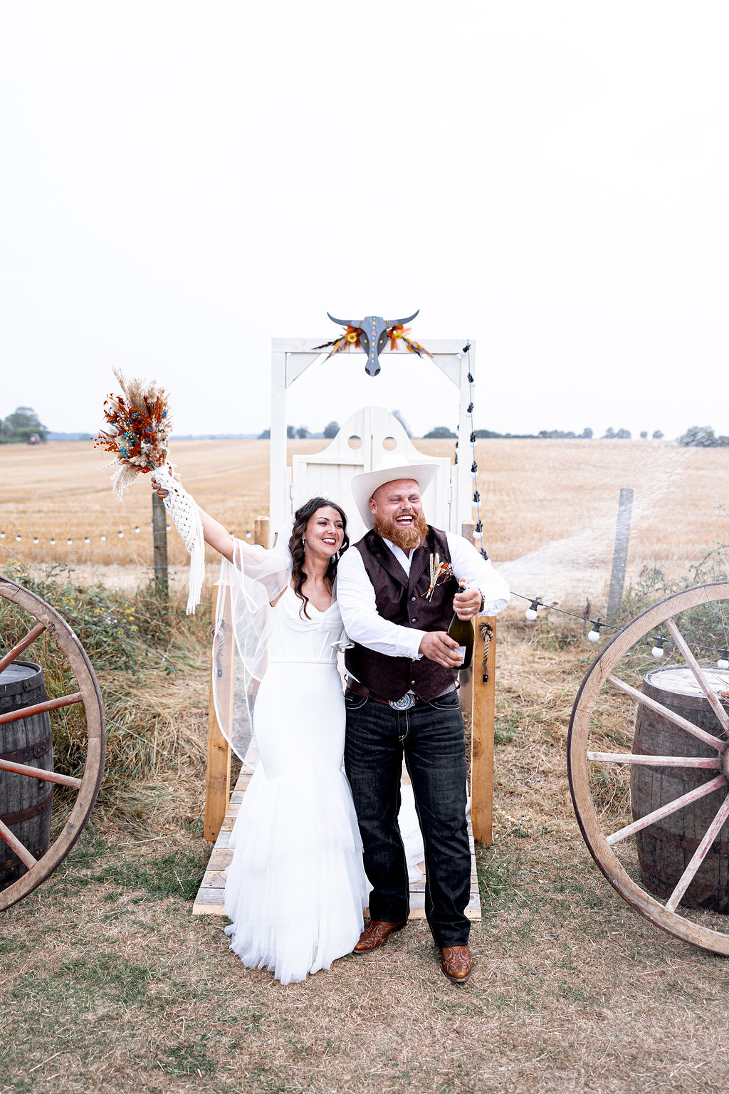 bride and groom stand in front of DIY western saloon doors spraying champagne. The bride holds a dried flower bouquet with macrame bouquet wrap