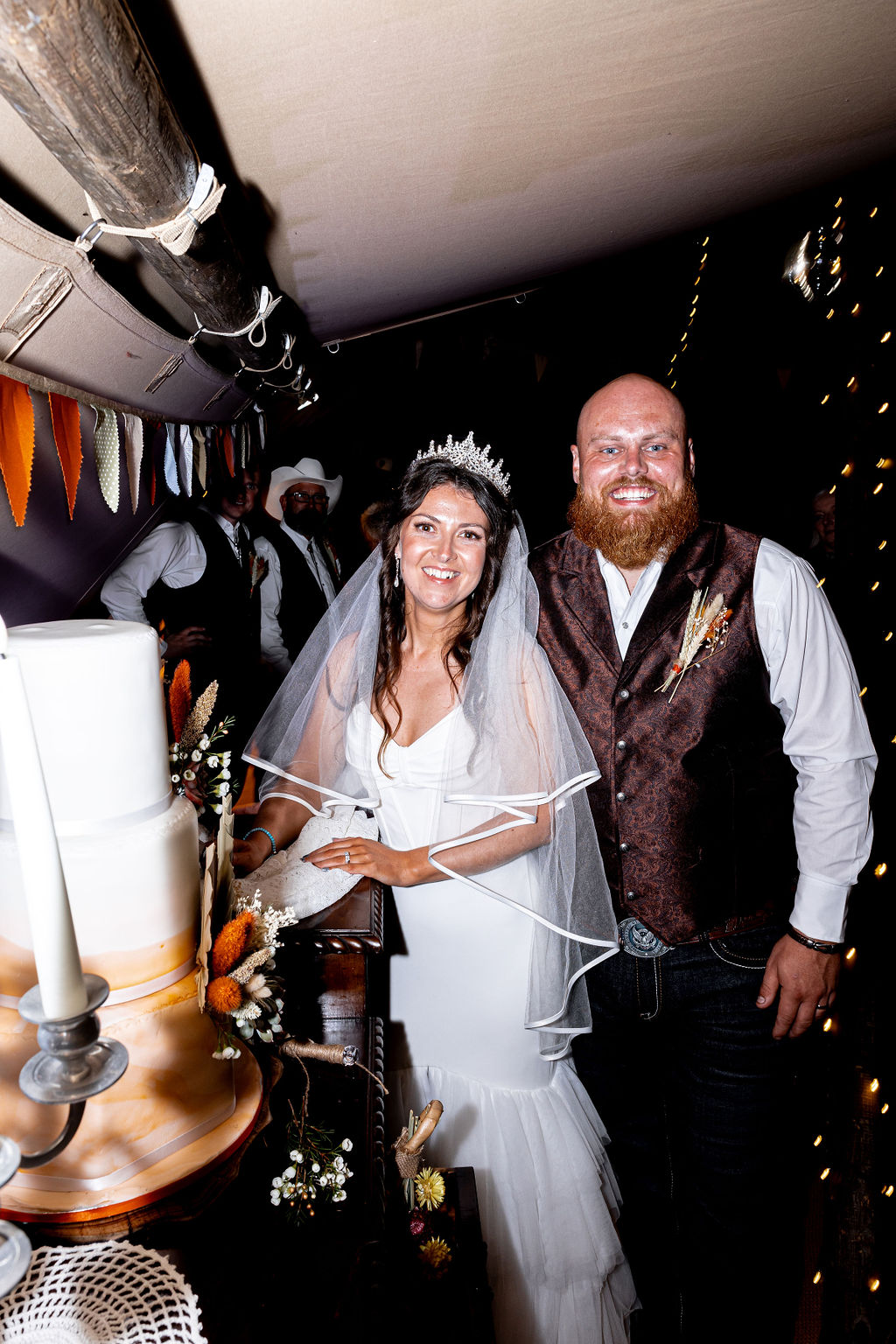 bride wearing ribbon trimmed veil and groom wearing western style waistcoat cutting their wedding cake