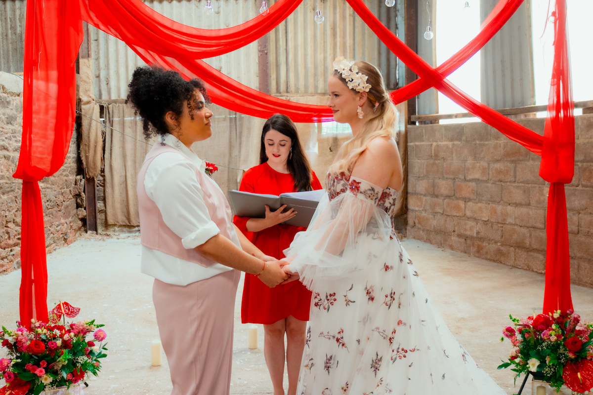 bride couple hold hands in front of wedding celebrant with red drape background