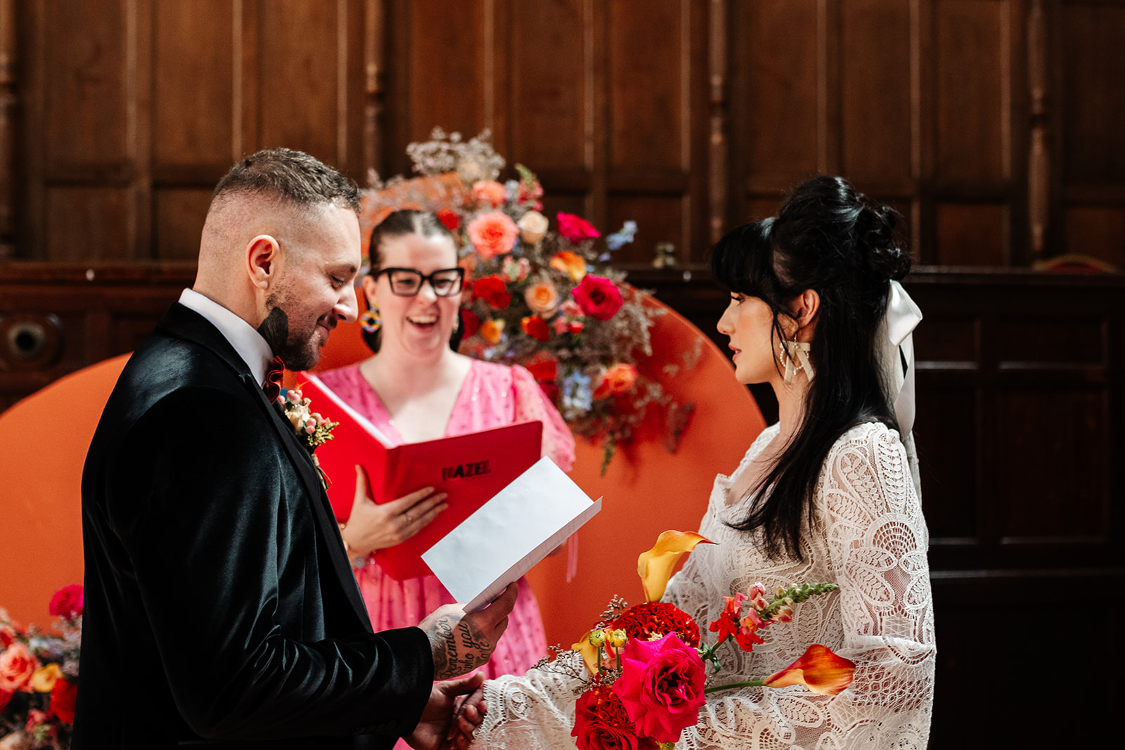 bride and groom stood holding hands while wedding celebrant laughs in the background