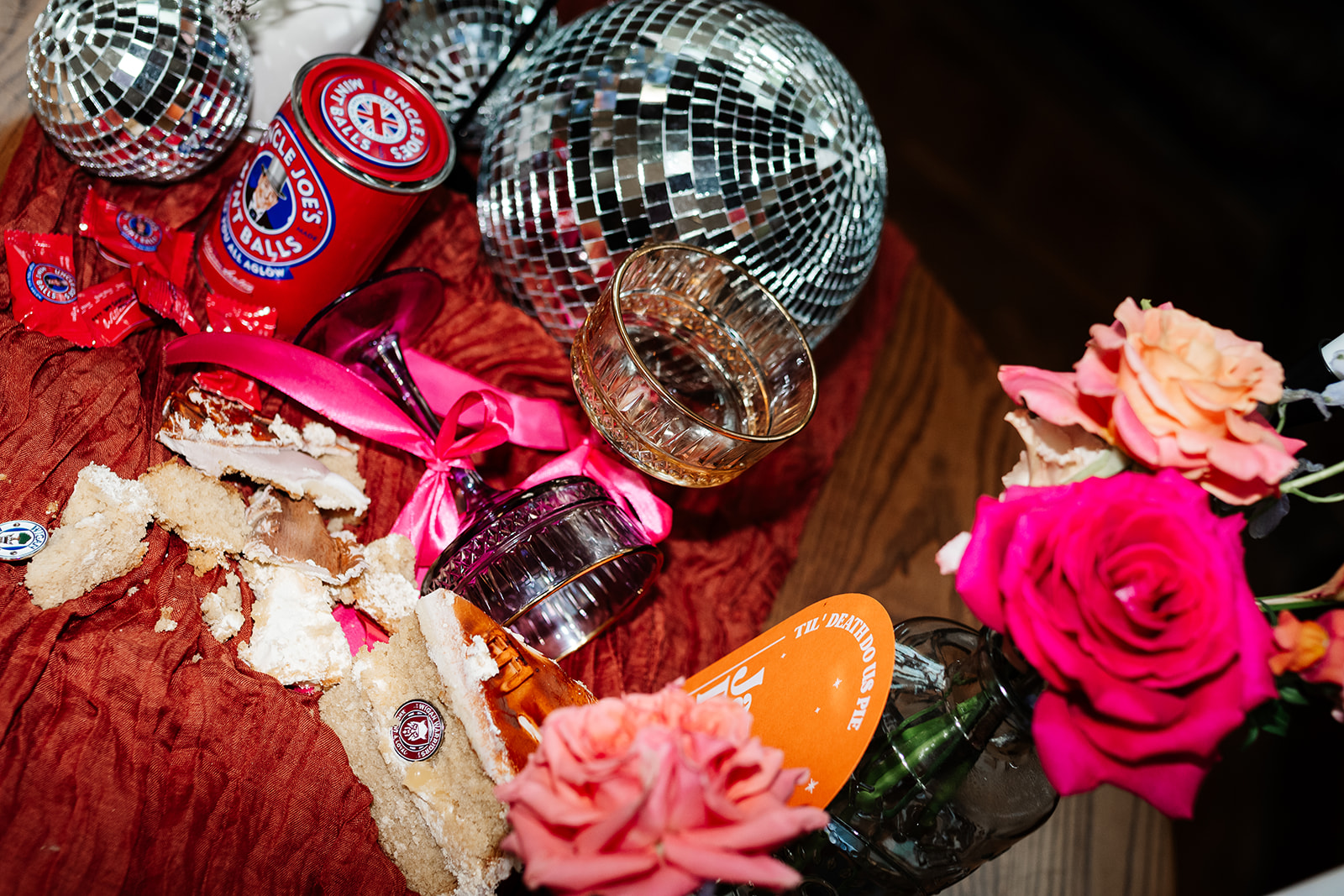 wedding flat lay of cake crumbs, champagne glass, uncle joes mint balls, disco balls and bright roses.