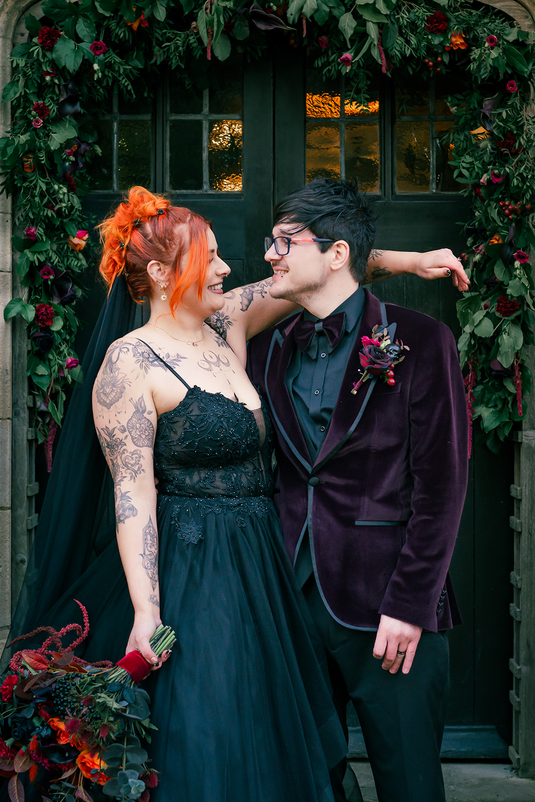 Bride with orange hair looks at the groom who wears a dark purple velvet suit jacket as part of a Halloween Wedding day.