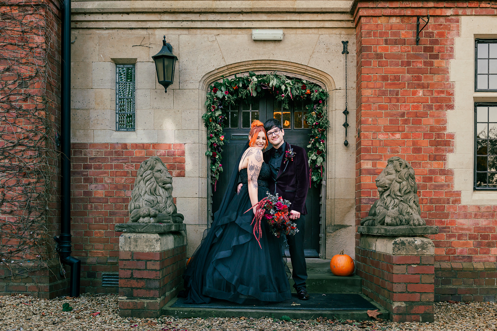Newely wed couple portrait outside of the wedding venue. The entrance has a deep green foliage with orange and deep red garland over it. A pumpkin sits on the stairs