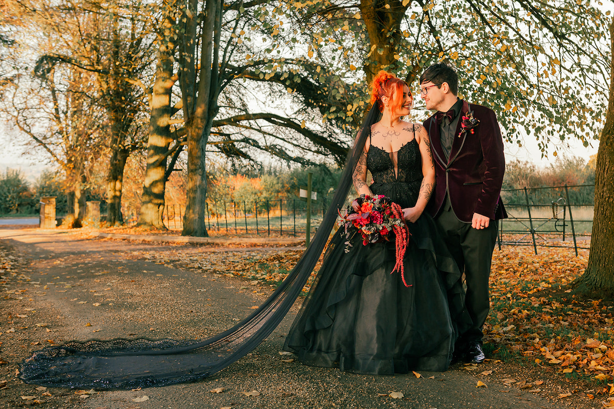 Newly wed couple stood in warm Autumn light - Bride wears black wedding dress with statement black veil and the groom wears plum velvet suit jacket with black shirt