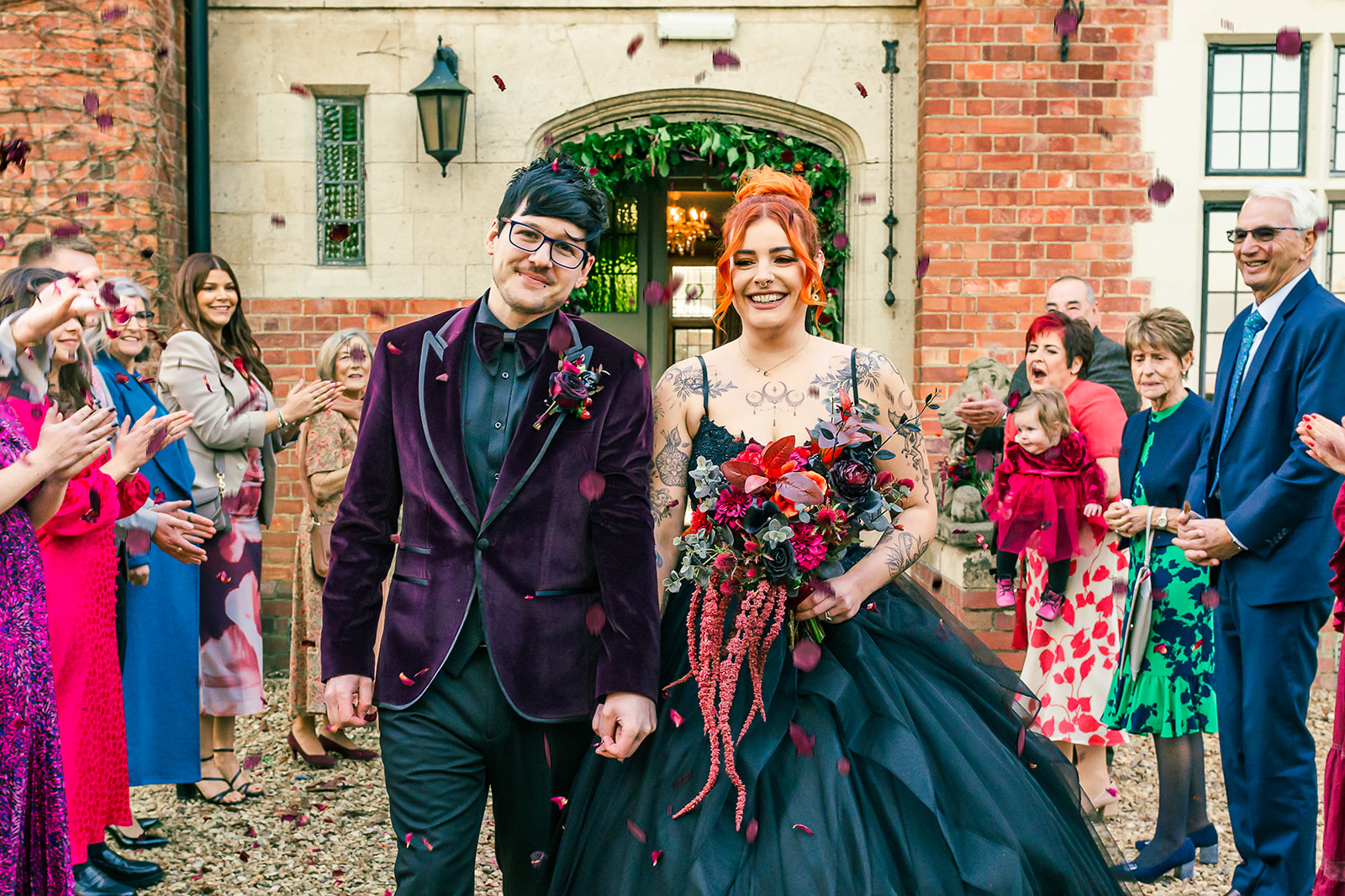 confetti shot of an alternative couple walking between guests wearing purple velvet suit jacket and black wedding dress.
