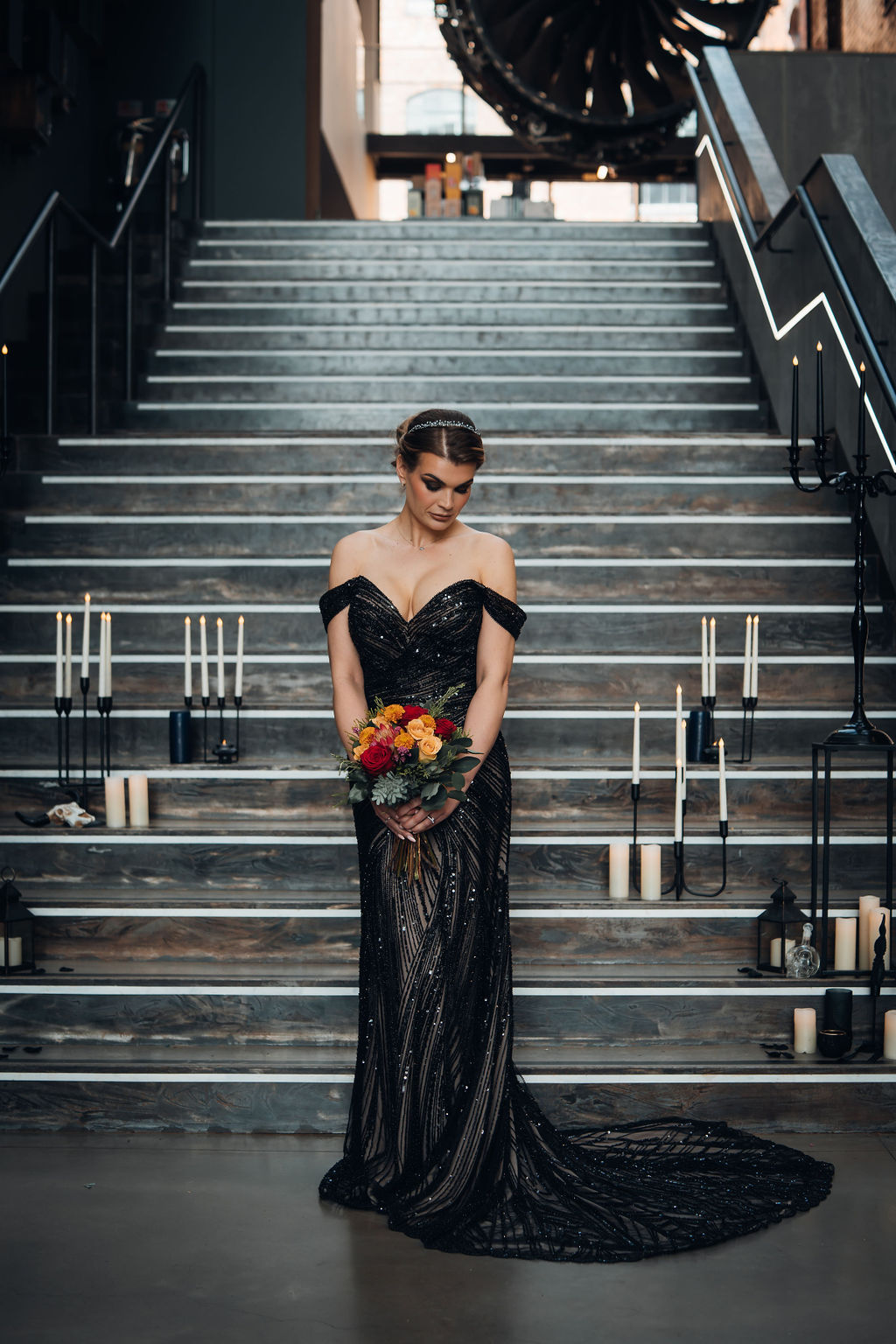 bridal portrait of a bride wearing a black beaded wedding dress stood in front of an industrial museum staircase covered with candles