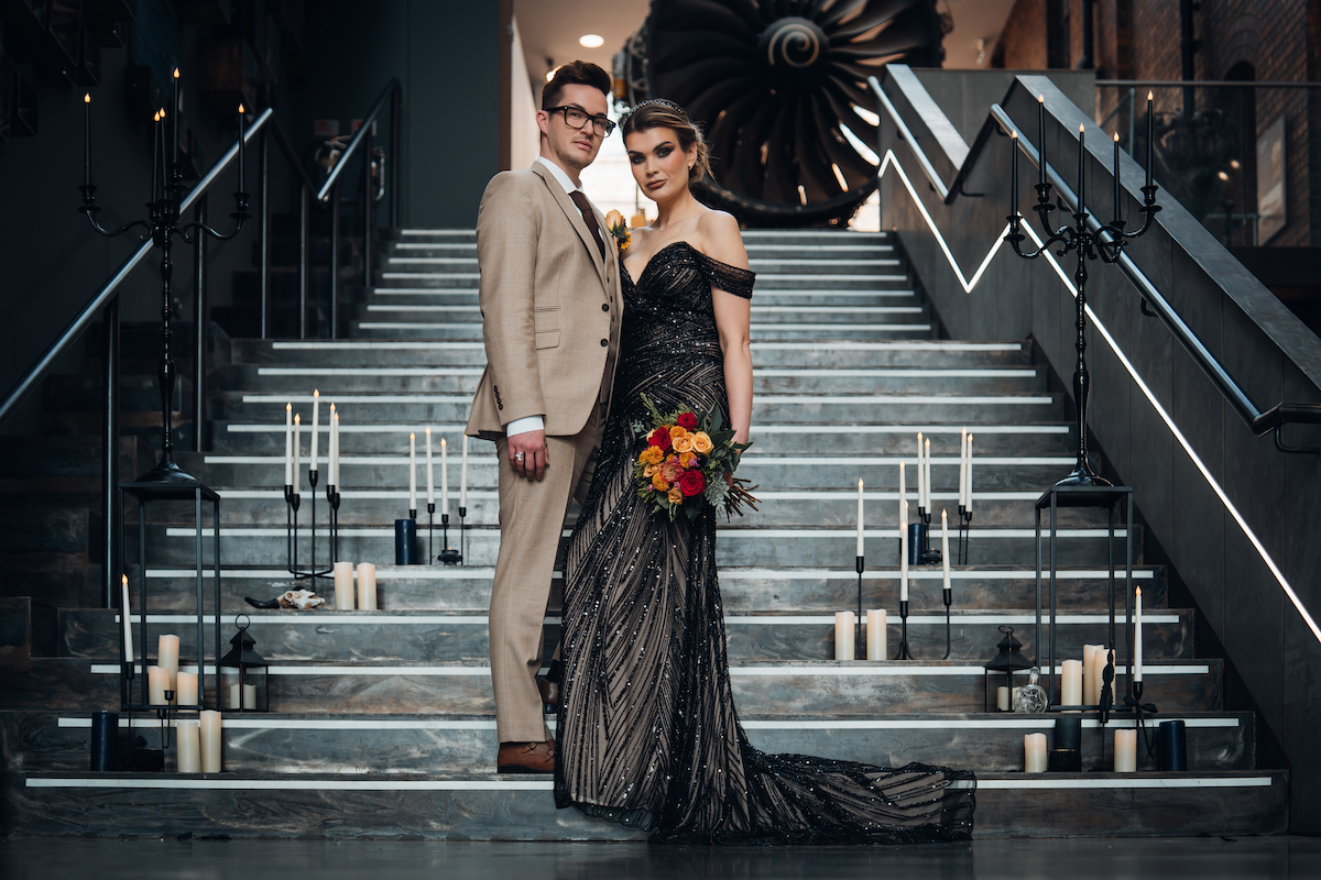bride dressed in a black wedding dress is stood with her groom on the staircase of an industrial museum surrounded with candles