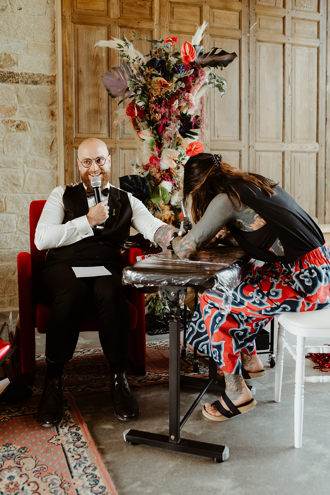 Groom talking in a microphone as a tattoo artist gives him a tattoo as part of the wedding ceremony