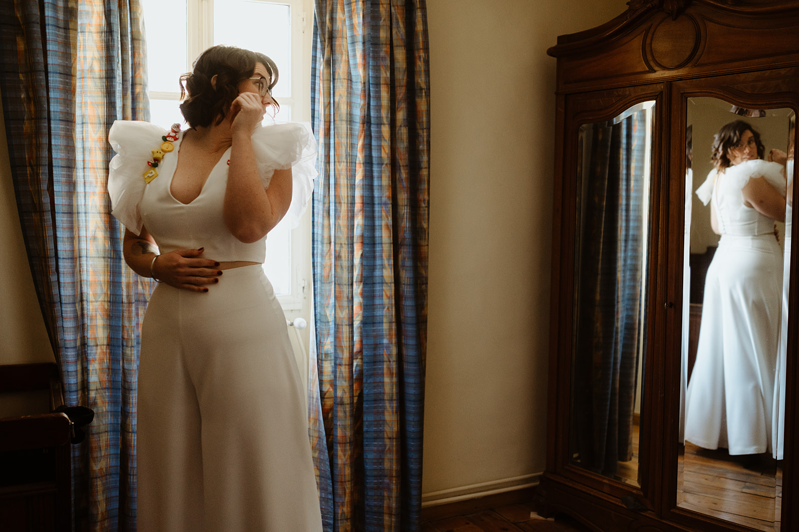 bride looking at herself in the wardrobe mirror. she wears an unconventional white two piece trouser outfit with ruffled shoulders