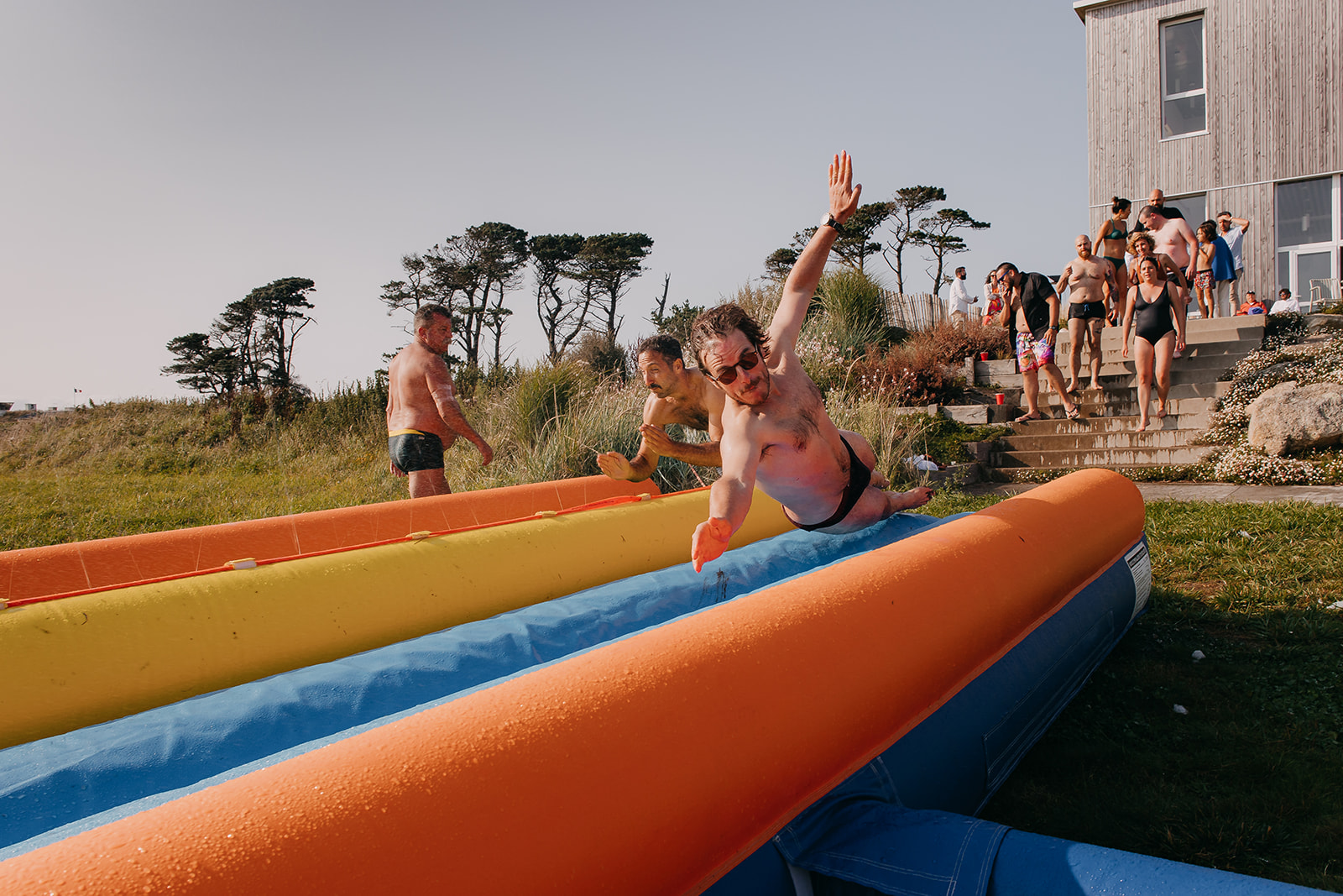 guests line up to take part in the slip and side at an unconventional french wedding. one guest is leaping through the air about to slide down.