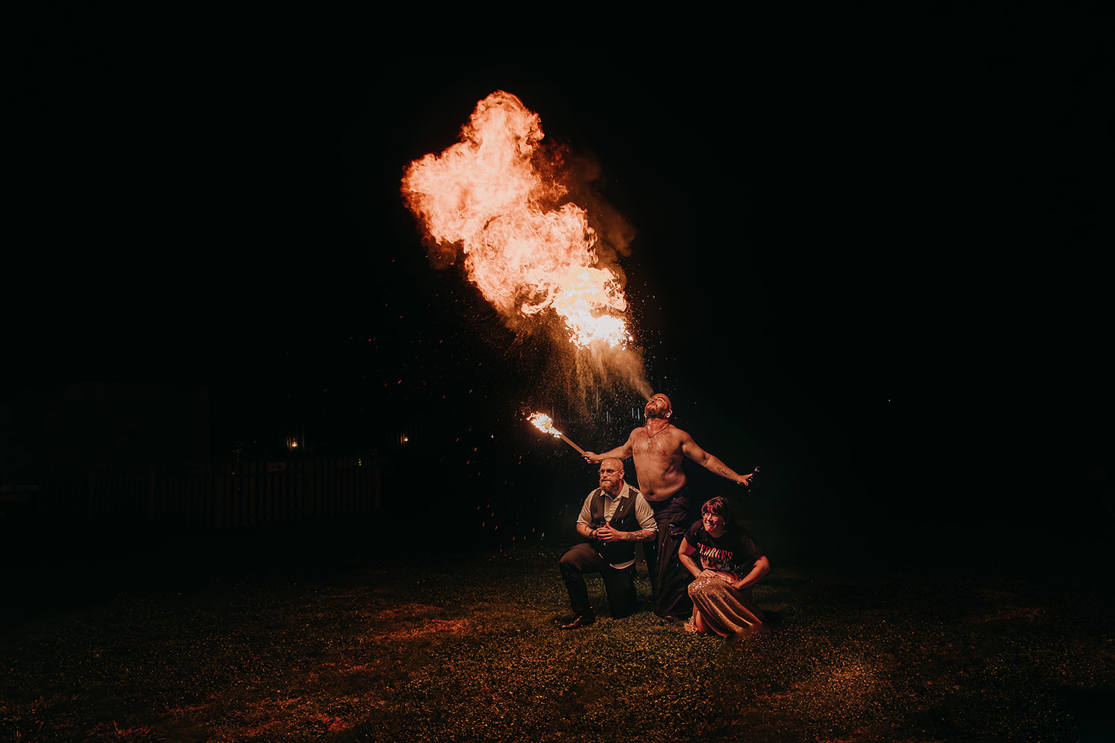 bride and groom sit under a fire breather who is breathing out a huge plume of fire at their unconventional wedding