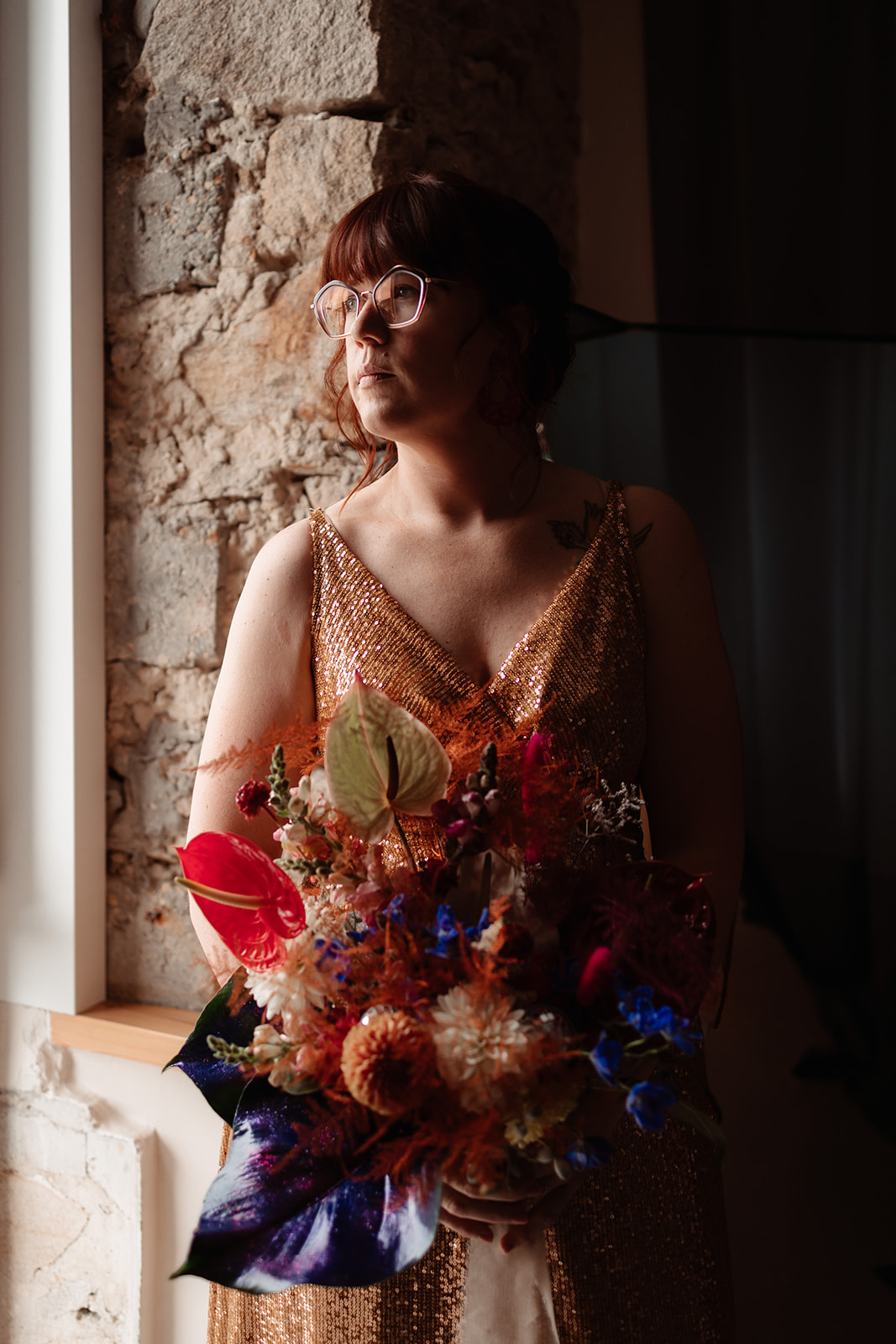 bridal portrait - the bride stands in hte window wearing a gold wedding dress and glasses. She holds a wedding bouquet made of red, blue and orange flowers