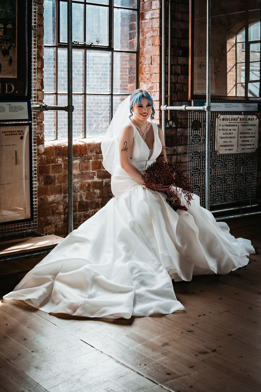 Bridal portrait of alternative bride with tattoos and blue hair sat in front of a window surrounded by metal shelving within an industrial museum