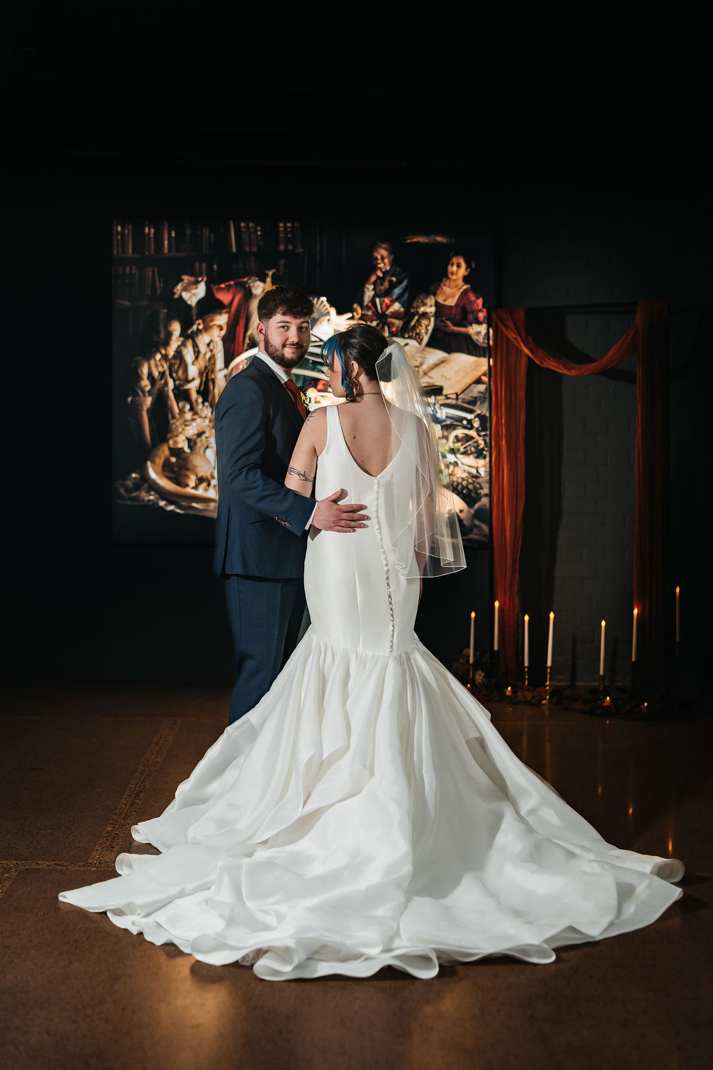 Bride and groom stand in front of a Joseph Wright painting lit by candlelight as part of their industrial wedding. The shot shows the deep V of the back of the brides dress and layered tulle mermaid train