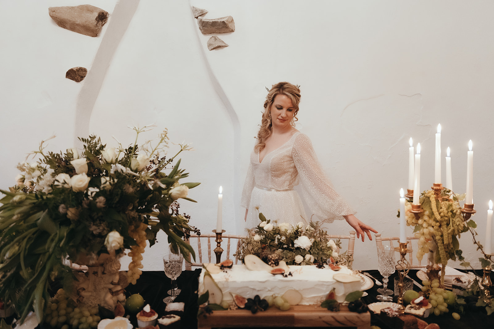 bride walks behind the chairs of her wedding breakfast table admiring candelabras, urns of flowers and grapes and rectangular wedding cake