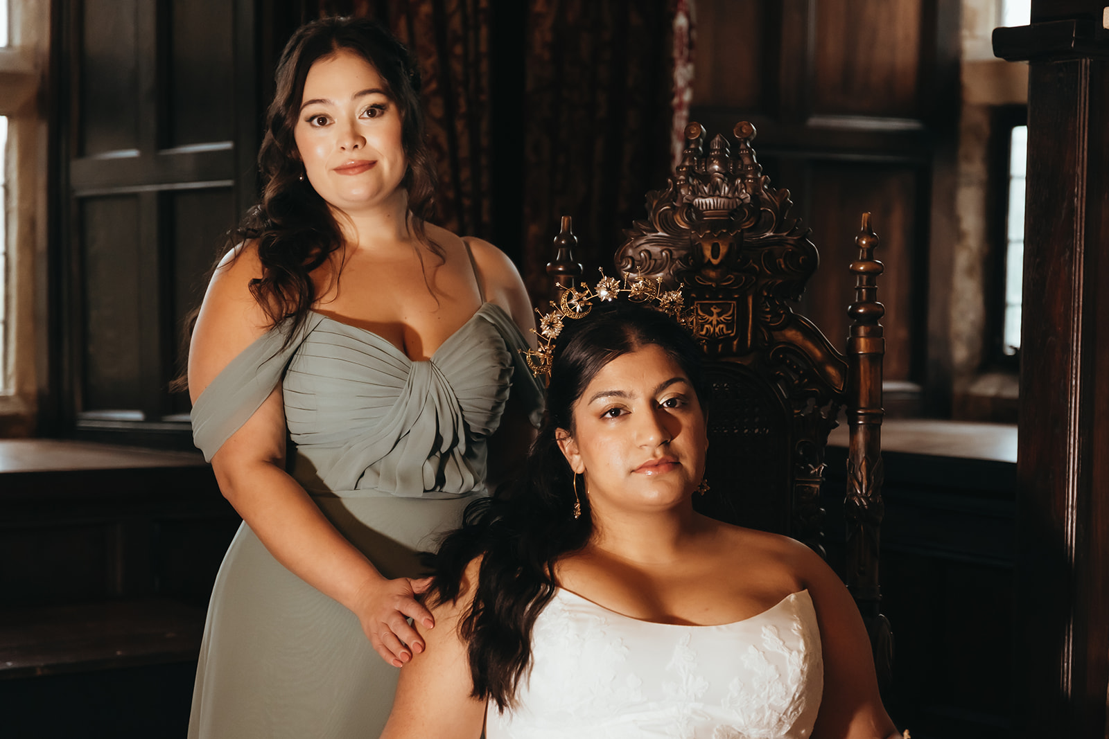 Bridal portrait with one bridesmaid. Bride wearing white and celestial crown sits in antique carved chair while bridesmaid in green stands at her side.