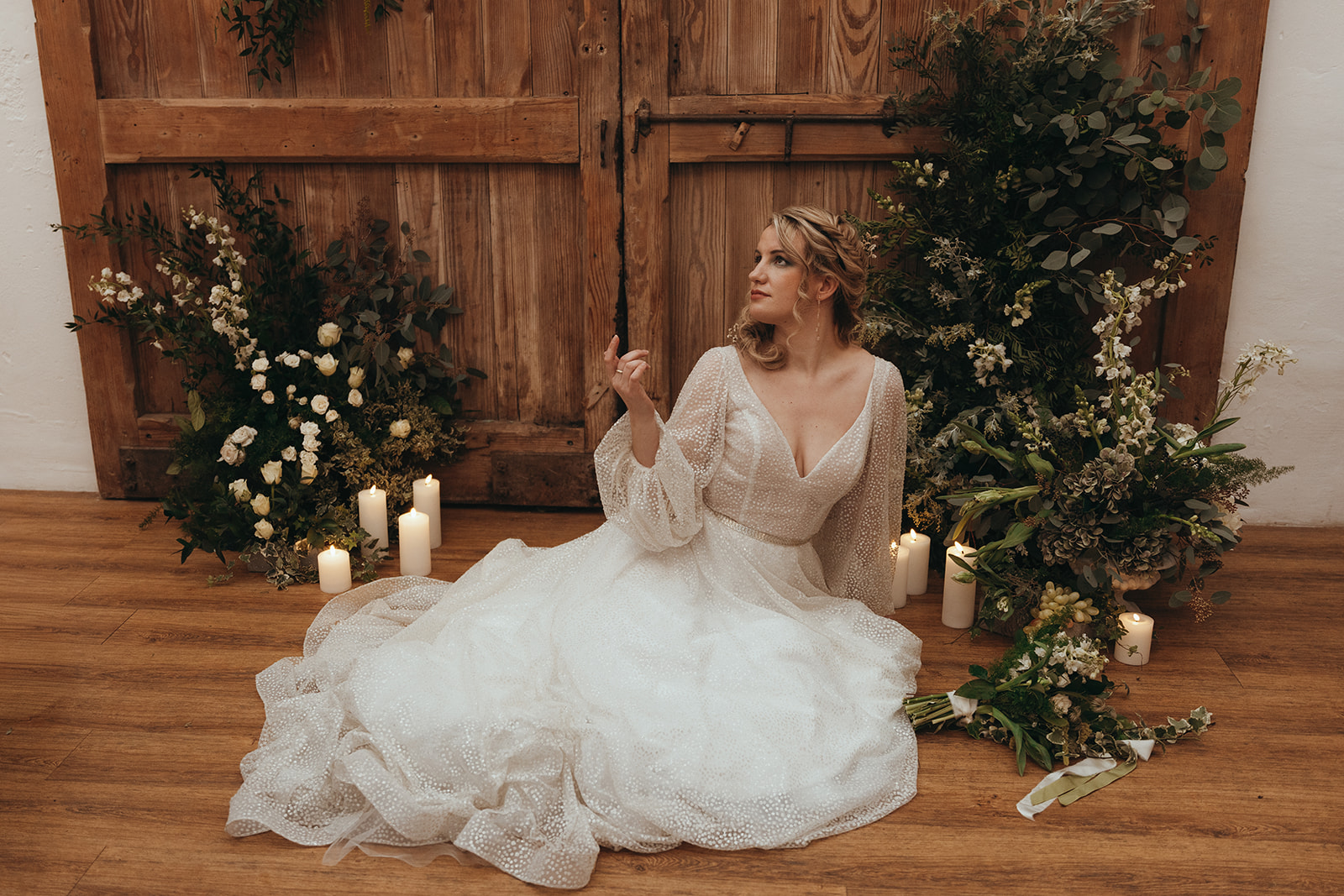 bride wearing a wedding dress with deep v and sleeves sits amongst asymmetrical floral arch and candles as part of a winter wedding