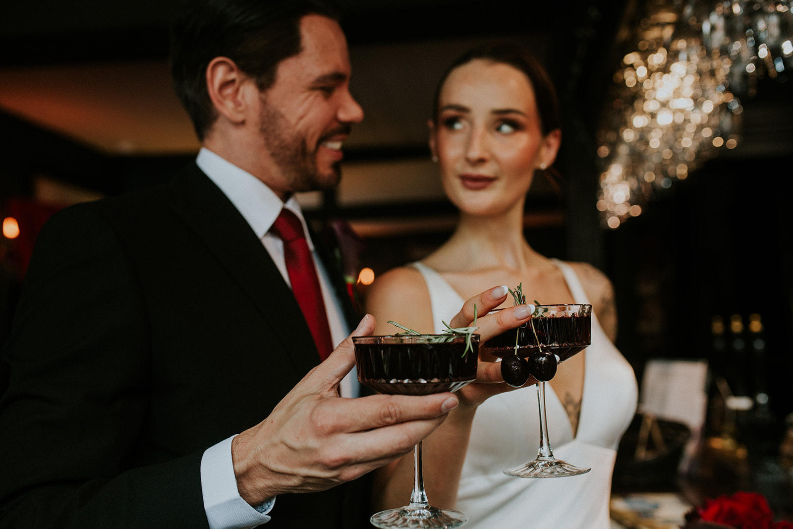 couple hold a deep red cherry cocktail as part of red winter wedding. the champagne glasses have cherries hanging from them