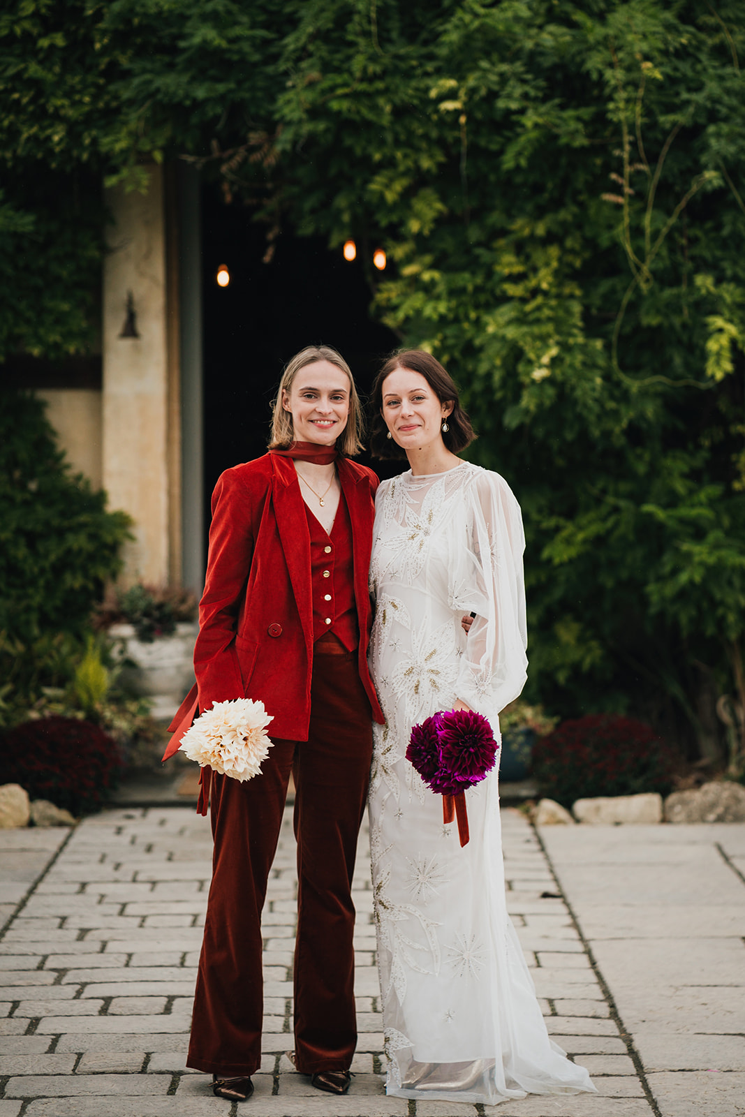 couple portrait where two brides stand together in front of their wedding venue. One wears white holding red flowers, the other wears red and hold white flowers