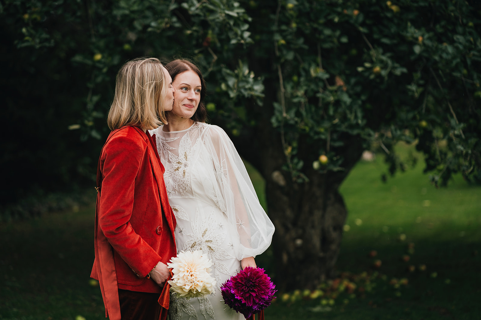 two brides - one wearing a white dress and the other a red suit hold home grown flower bouquets