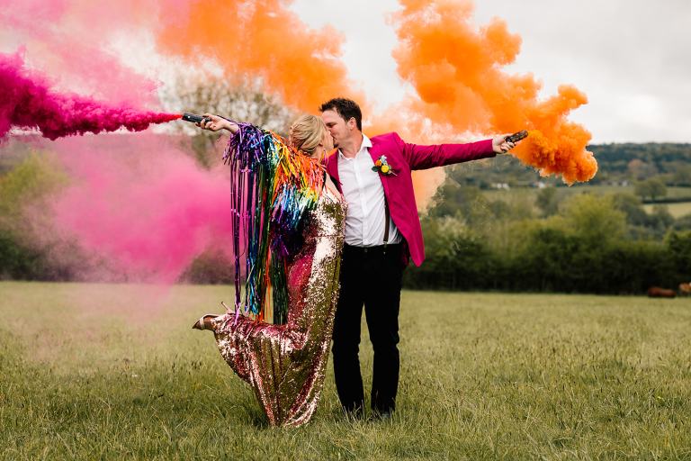 Alternative wedding fair by quirky weddings, at the mill farm in cambridge. Newlywed couple stand in a field with colourful smoke bombs