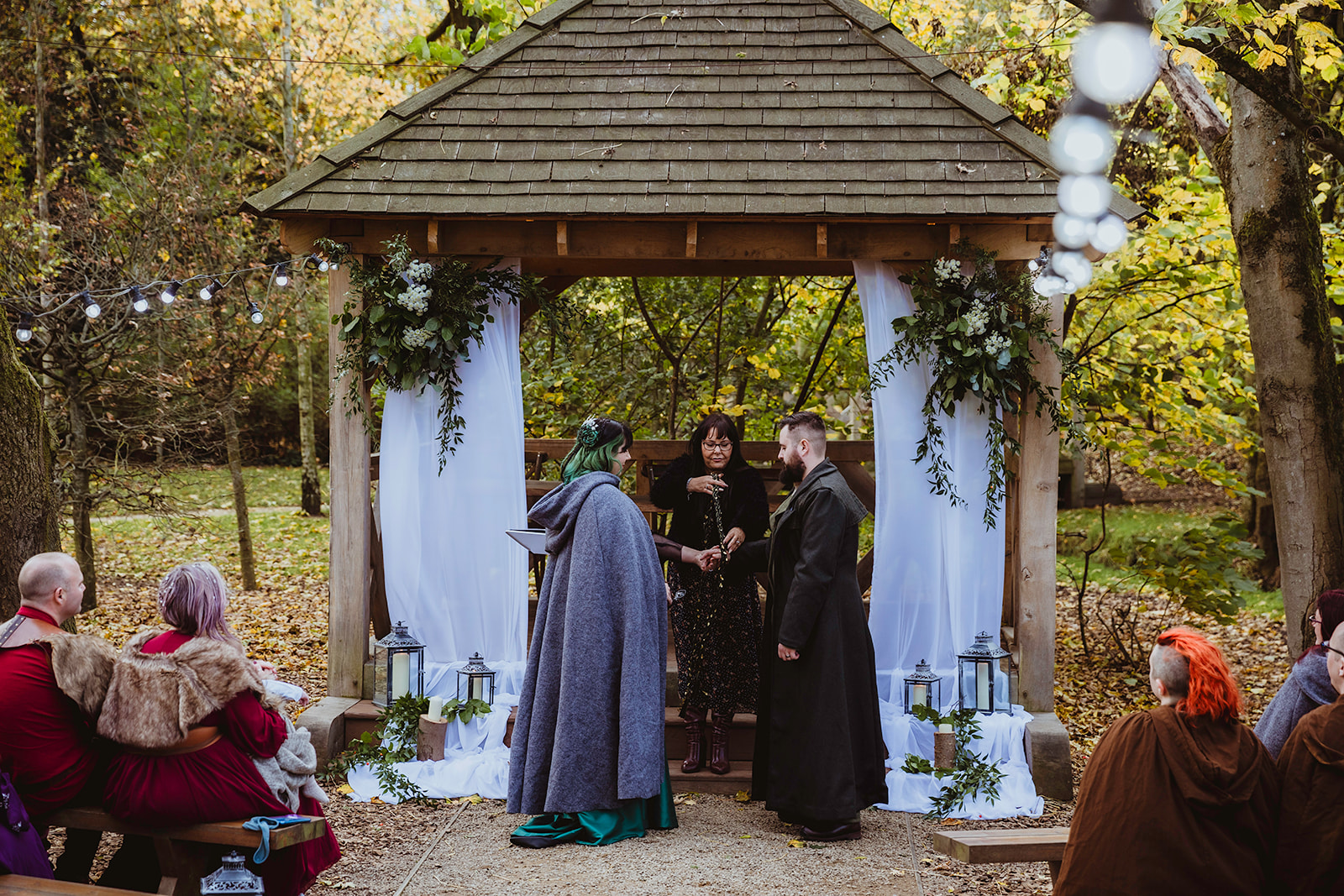 bride and groom stood under the wooden structure in a woodland wedding venue ready for handfasting ceremony