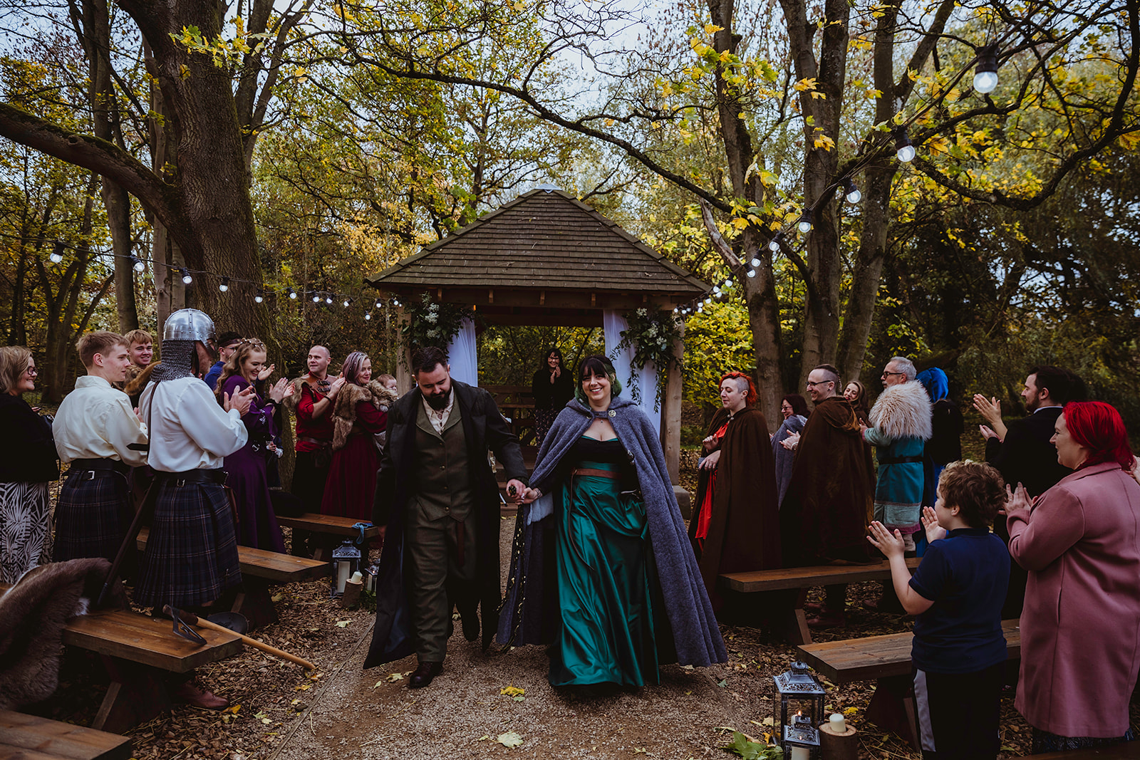 bride and groom walk down the woodland aisle with wedding guests wearing Viking inspired outfits either side.