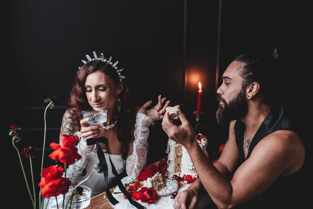 bride drinks champagne and groom eats wedding cake both surrounded with deep red candles and flowers as part of their dark romance couple photoshoot