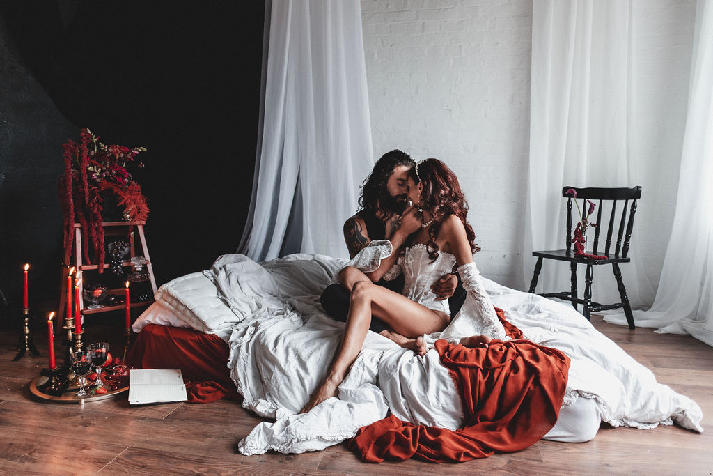 couple embrace on a floor bed surrounded with deep red flowers, candles and fruit as part of a couples boudoir session