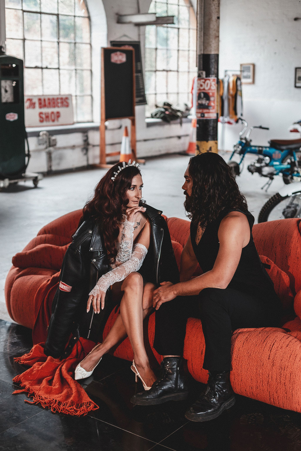 couple sit on a large orange sofa in the middle of a motorbike studio for alternative wedding portraits