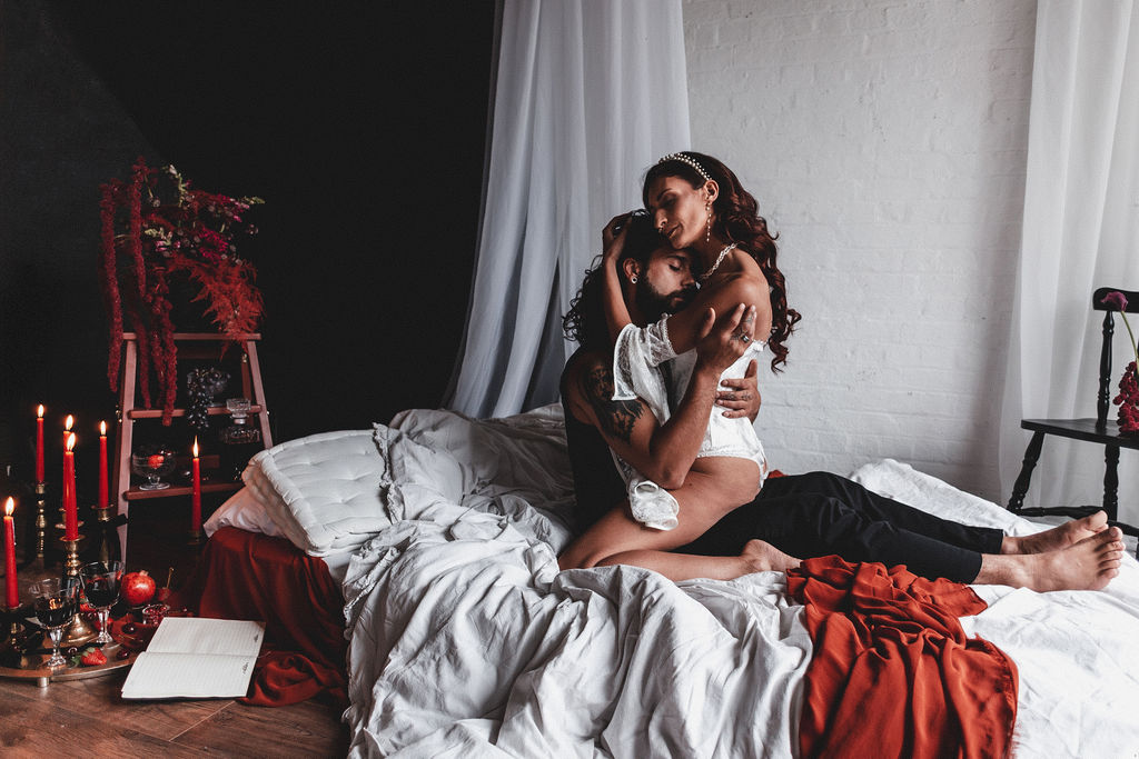 couple embrace on a floor bed surrounded with deep red flowers, candles and fruit as part of a couples boudoir session