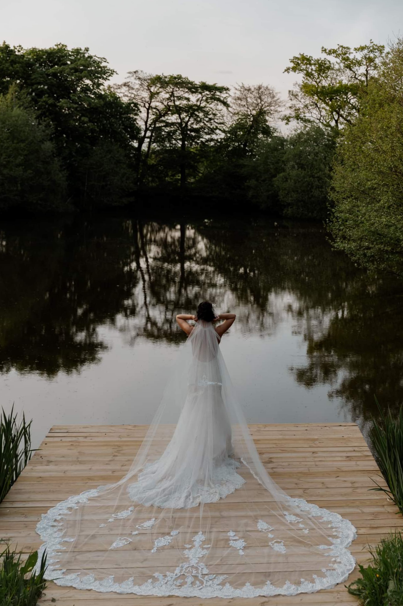 Bride stood on a jetty by a lake with her lace wedding veil by Lisa Carrington Couture out behind he