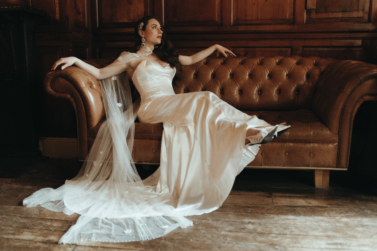 Bride wearing chic wedding dress and Hollywood waves at on a brown sofa. She wears a floral cape as part of her outfit.