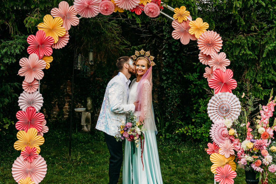 bride and groom stand under a paper flower ceremony arch at their festival wedding
