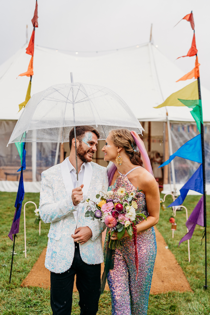 Bride and groom waring sequins walk arm in arm holding umbrellas as part of their outdoor glitter festival wedding