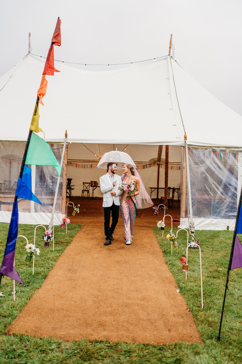 Bride and groom waring sequins walk arm in arm holding an umbrella in front of their wedding marquee