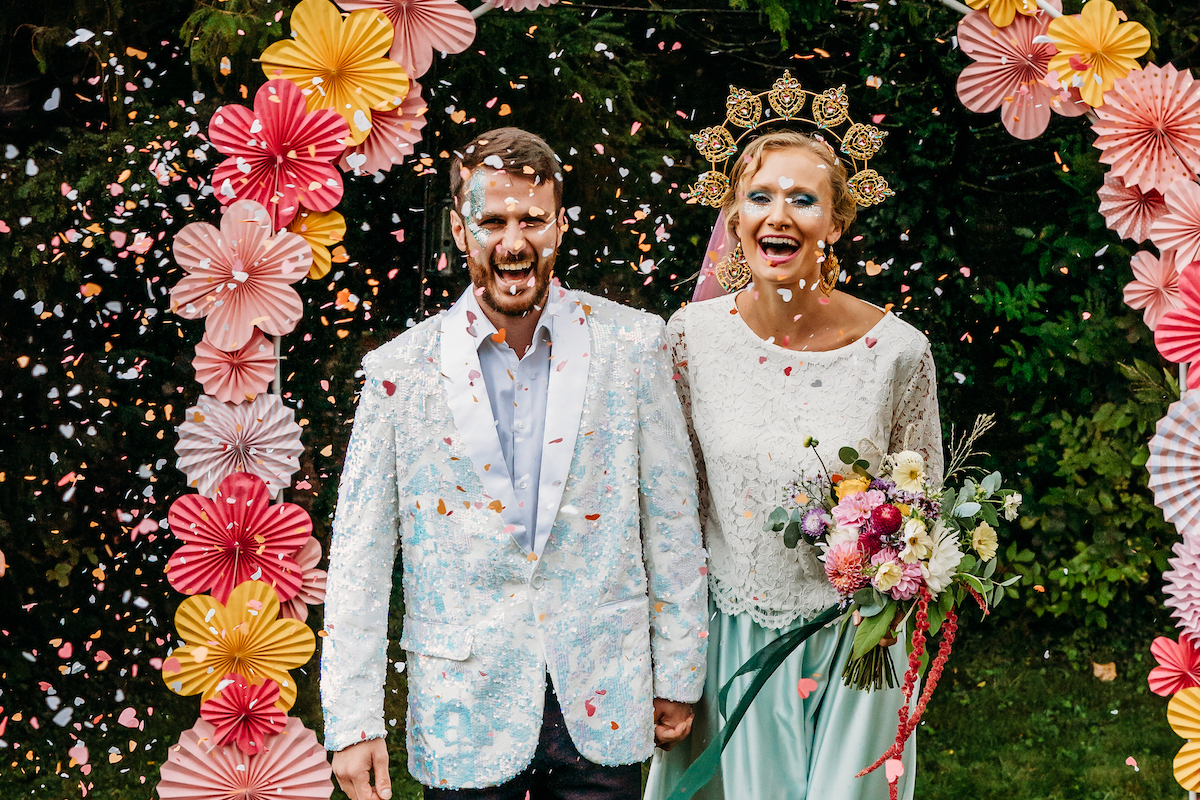 Bride and groom surrounded with confetti with a colourful paper flower wedding ceremony arch in the background