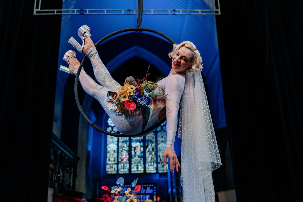 bride wearing iridescent jump suit and veil holding a bouquet of vibrant flowers sat in an areal hoop