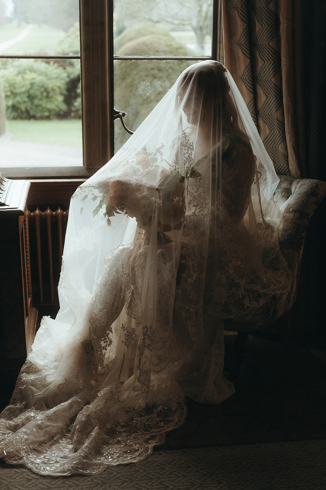moody bridal portrait where the bride is backlit, sat at a window with her veil over her.