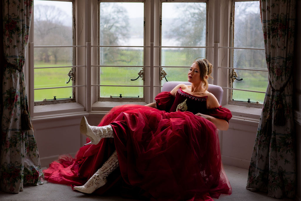 bride wearing a red wedding dress and lace knee high boots sat in the window of her castle wedding venue