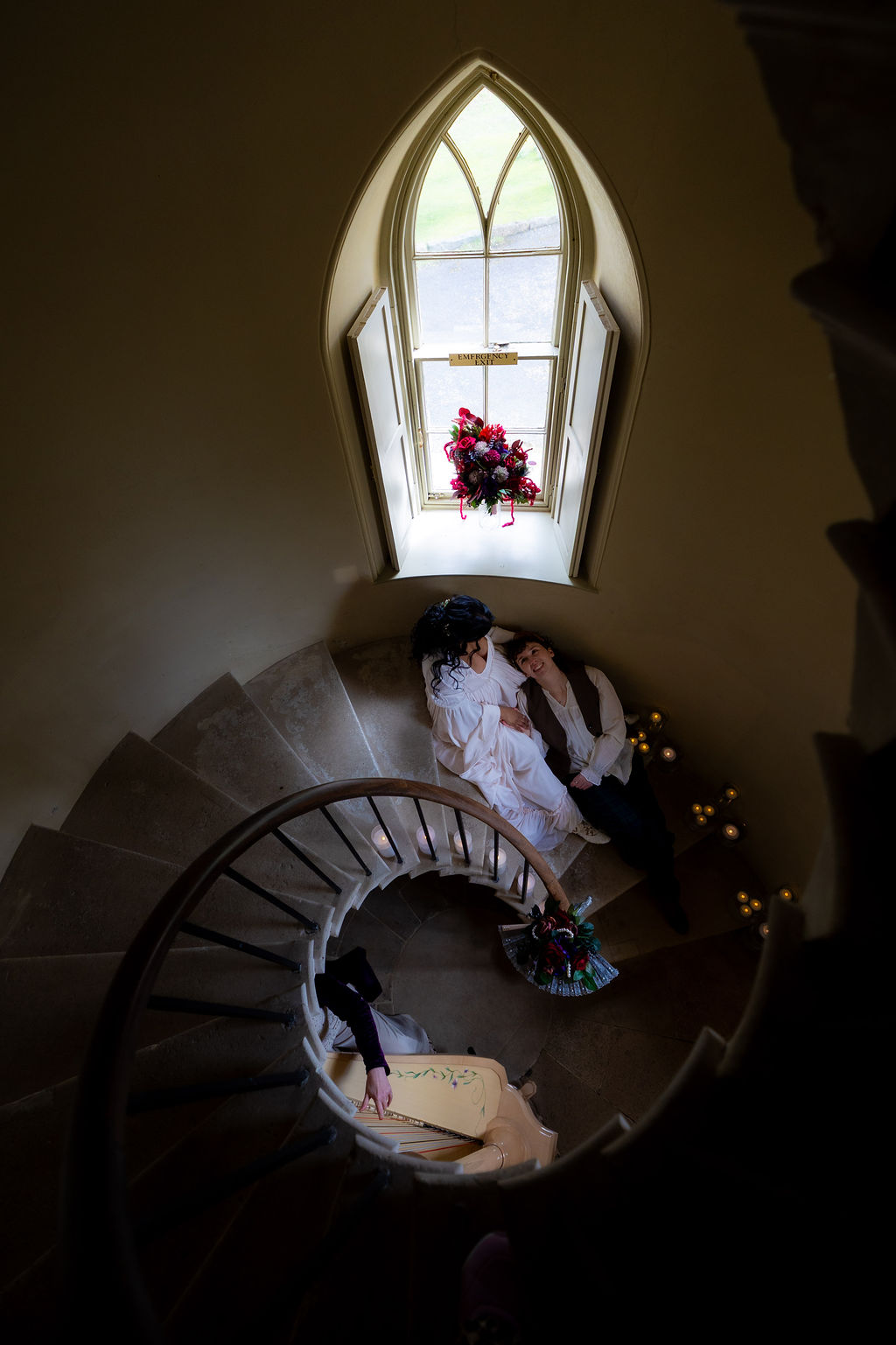 couple sat in candle lit circular stairwell of a historic Devon wedding venue with harp player below