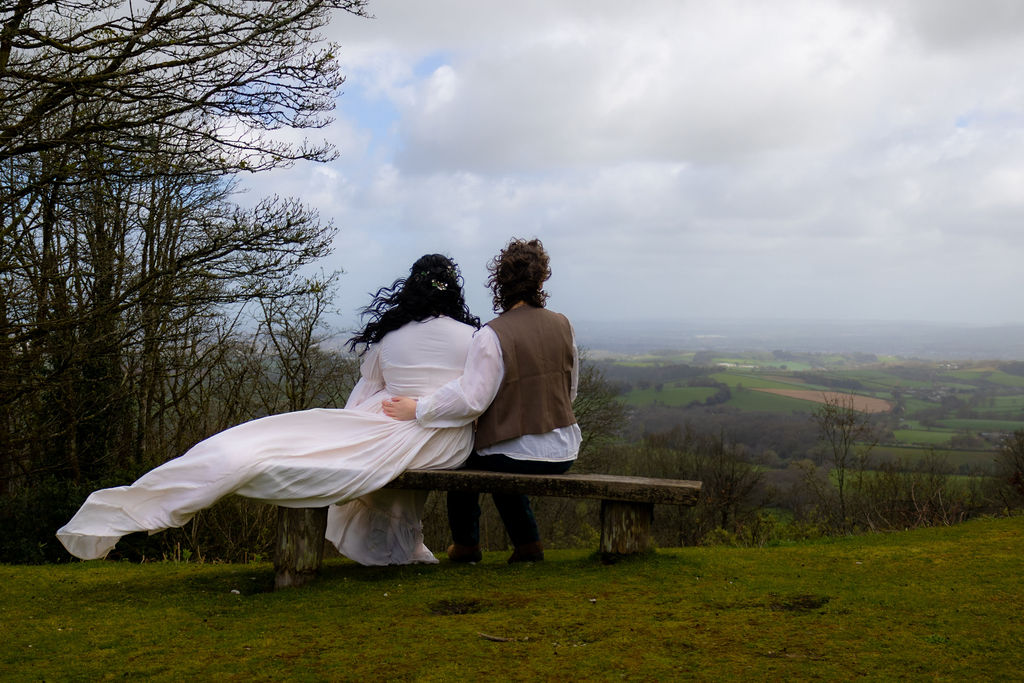 couple sitting on a bench looking at the Devon landscape with the wind blowing their historic style wedding dress